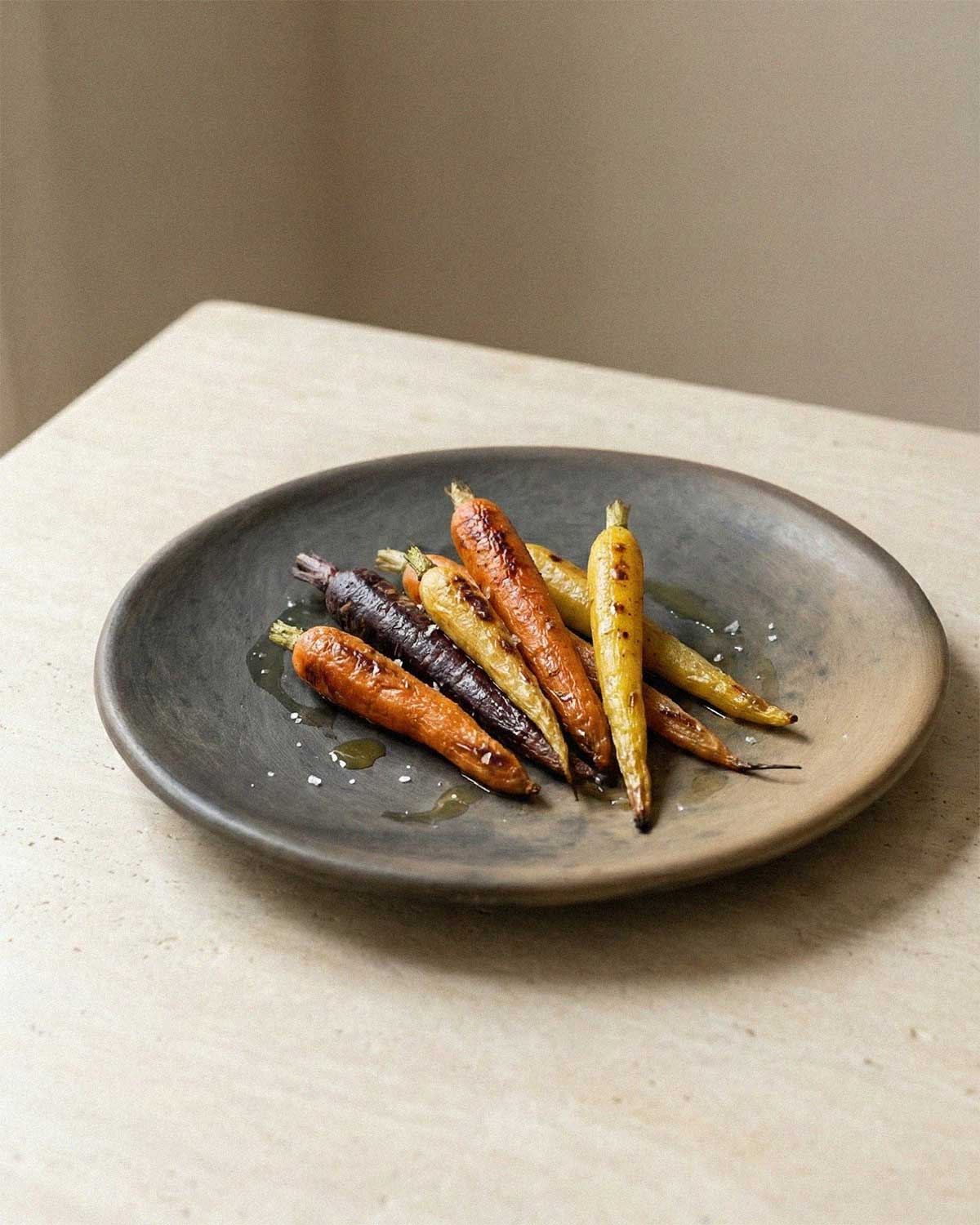 Angled view of the handmade Oaxacan Clay Dinner Plate showing its unique dark grey and brown finish, presenting a serving of roasted rainbow carrots drizzled with oil and coarse salt.
