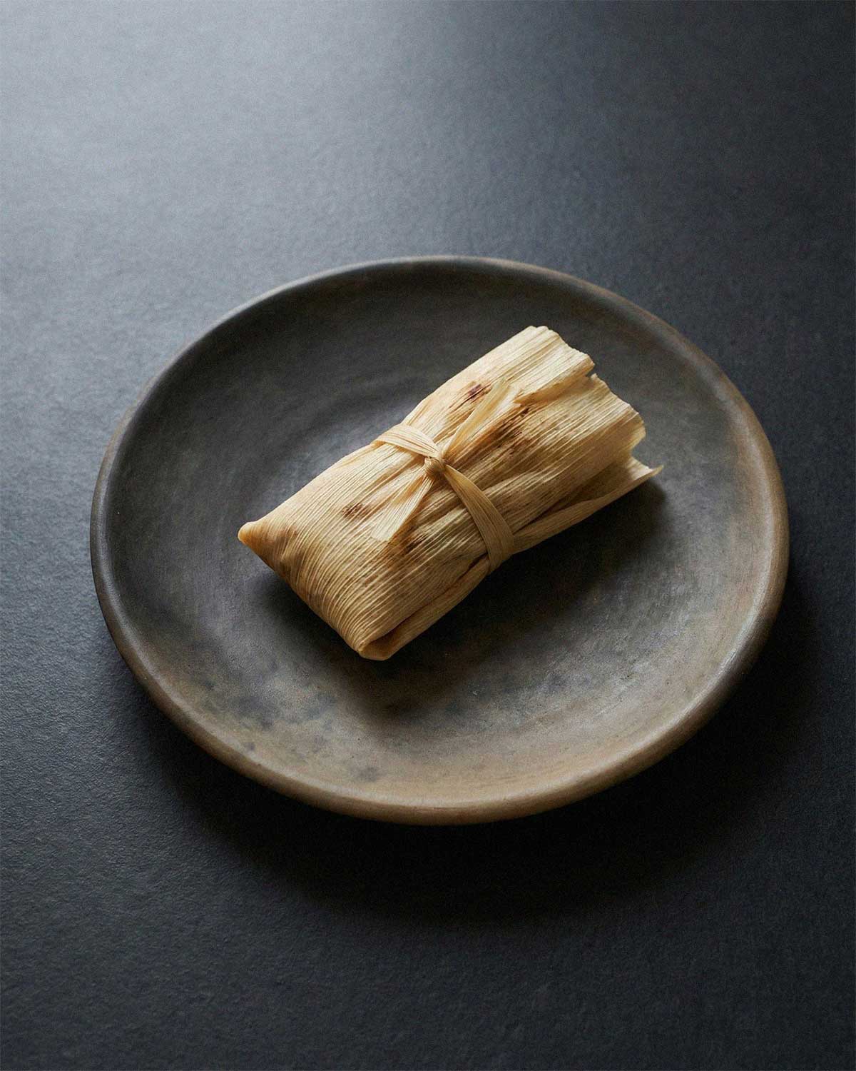 Top view of the handmade Oaxacan Clay Dinner Plate showing its dark, smoke-fired variations, holding a single corn husk-wrapped tamale tied with a husk strip.