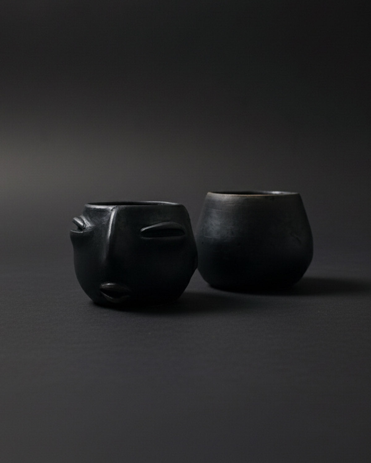 A pair of handmade Oaxacan clay cups on a dark surface, one with a carved face and one plain.